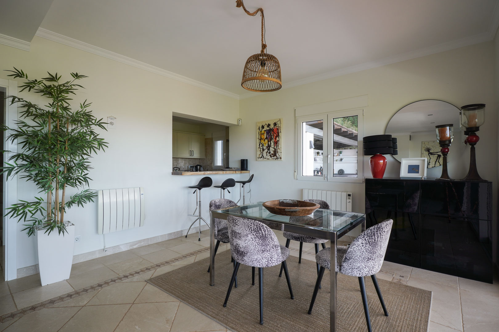 Comedor luminoso con mesa de cristal, sillas tapizadas y lámpara de fibras naturales, conectado a la cocina semiabierta mediante una barra con taburetes.