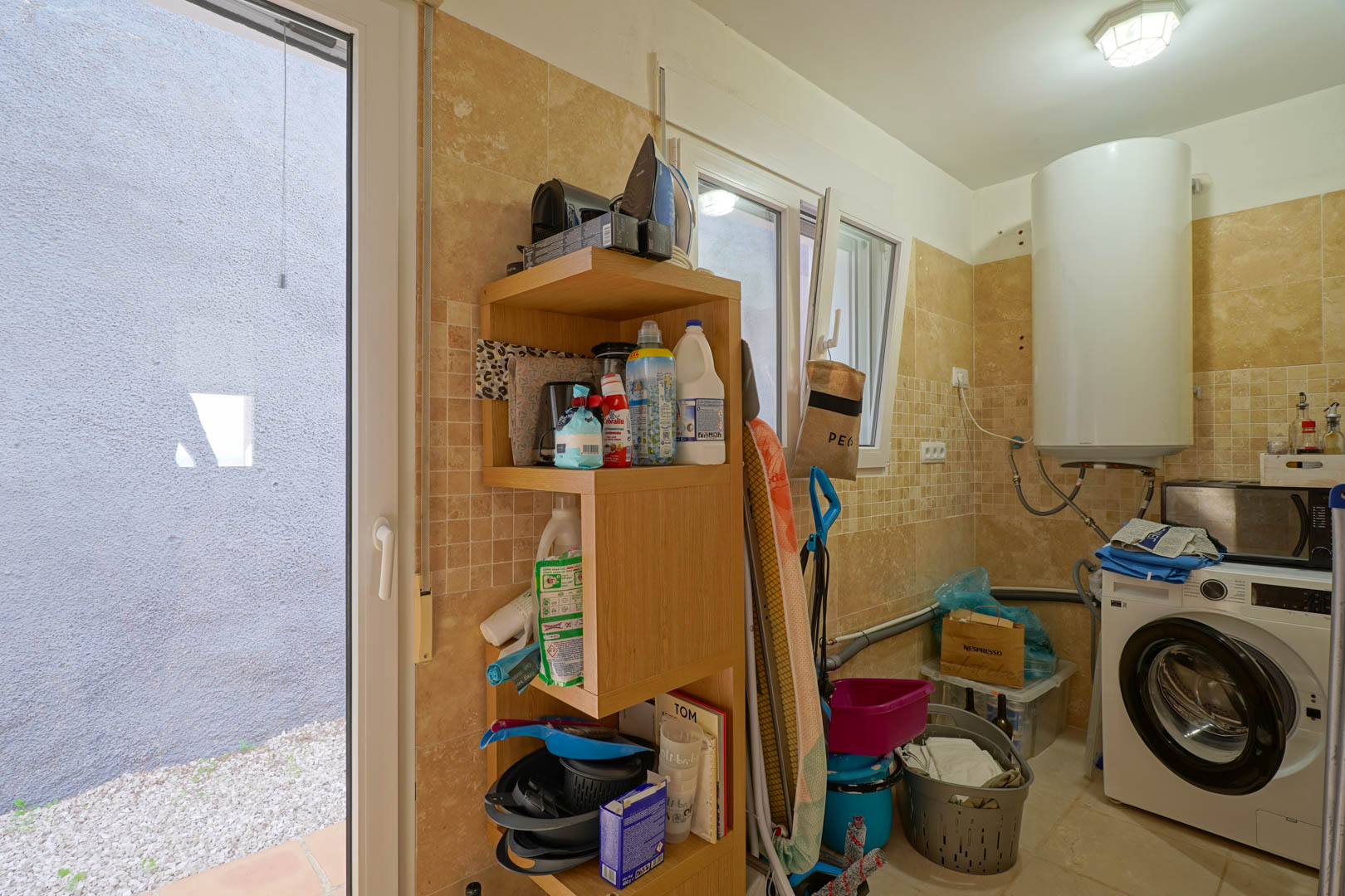 Cuarto de lavandería funcional con paredes alicatadas en tonos crema, equipado con termo eléctrico, lavadora, estantería de madera y acceso directo al exterior.