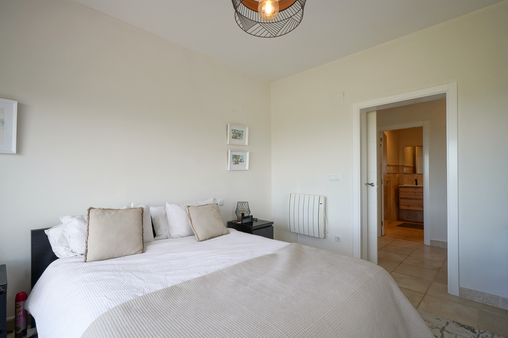 Dormitorio luminoso con suelos cerámicos, cama doble y acceso directo a baño en suite. Incluye radiador eléctrico y lámpara de techo con diseño de rejilla metálica.