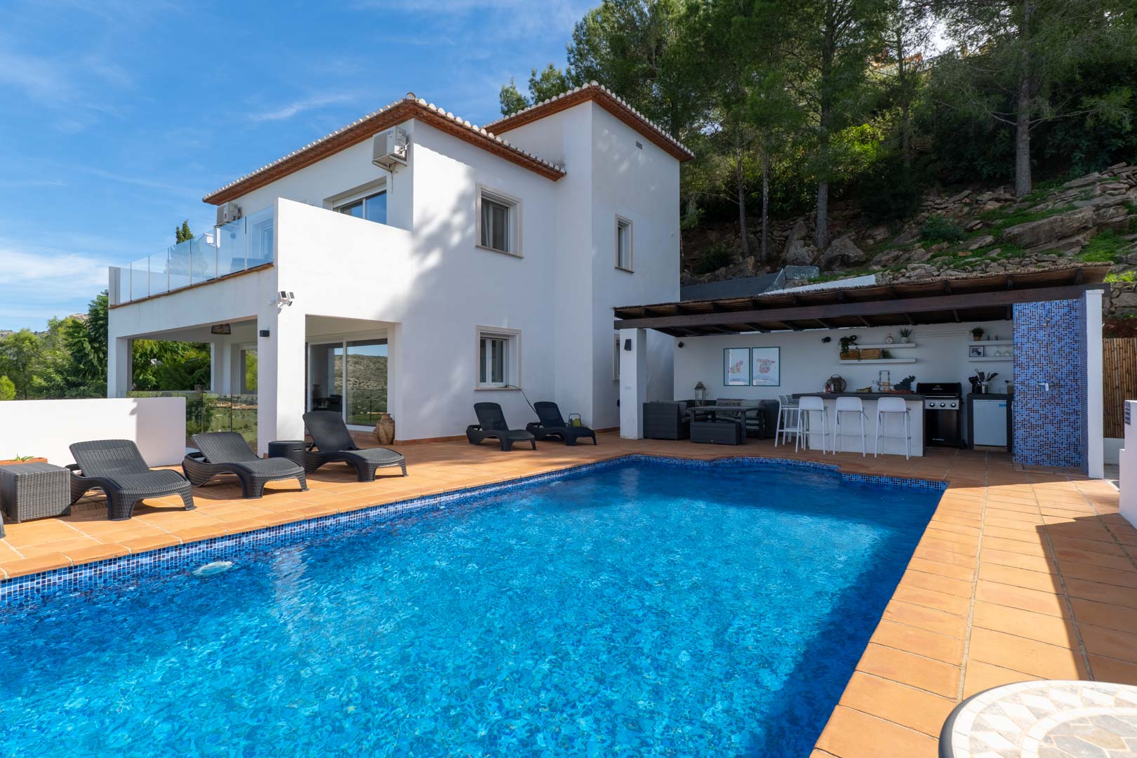 Exterior con piscina de gresite azul, terraza de terracota y cocina de verano equipada. La fachada blanca de estilo mediterráneo linda con una zona boscosa de pinos.