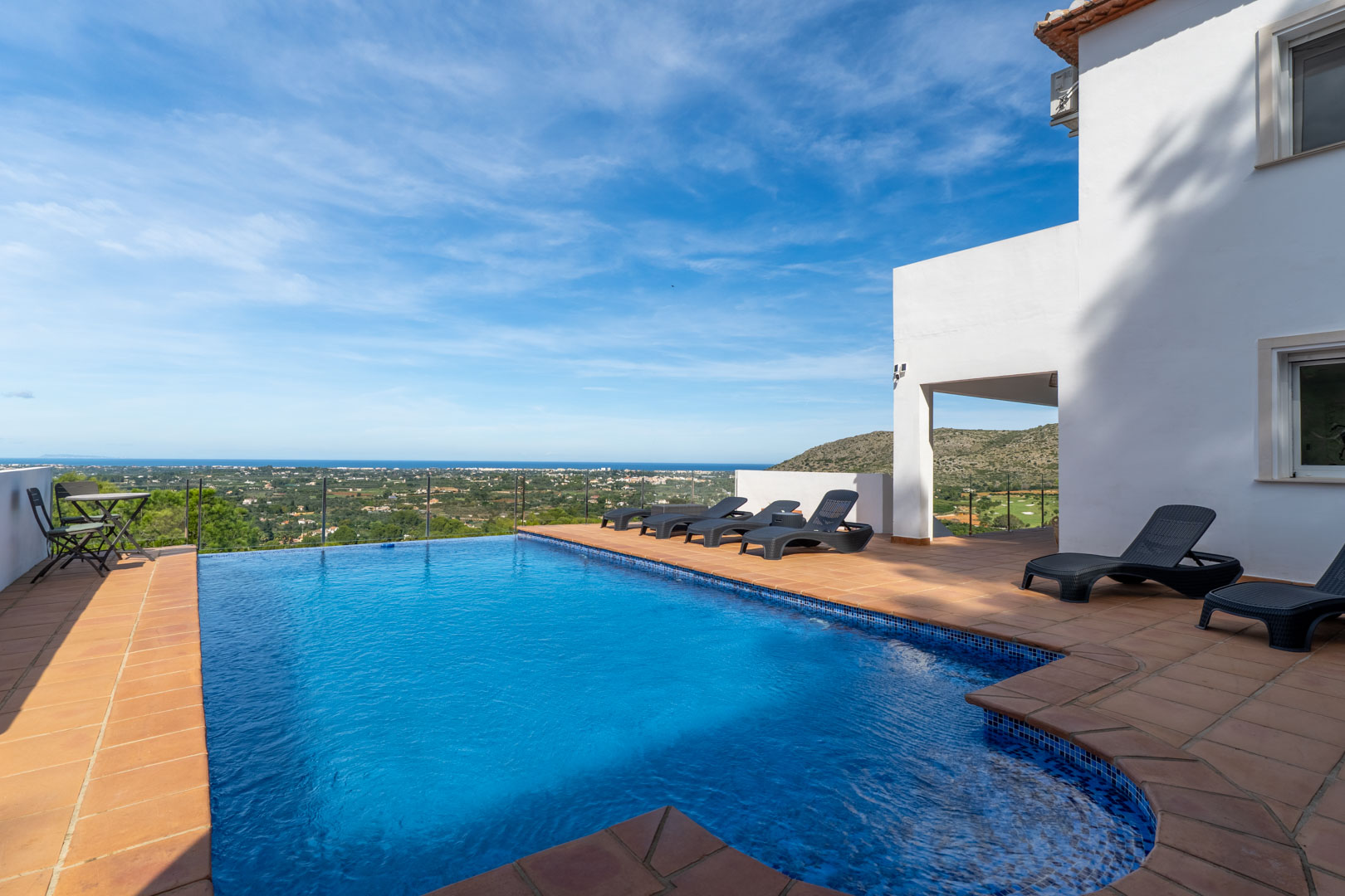 Piscina privada con terraza de baldosas de barro, tumbonas negras y barandilla de cristal con vistas panorámicas al mar, el valle y el campo de golf de La Sella.