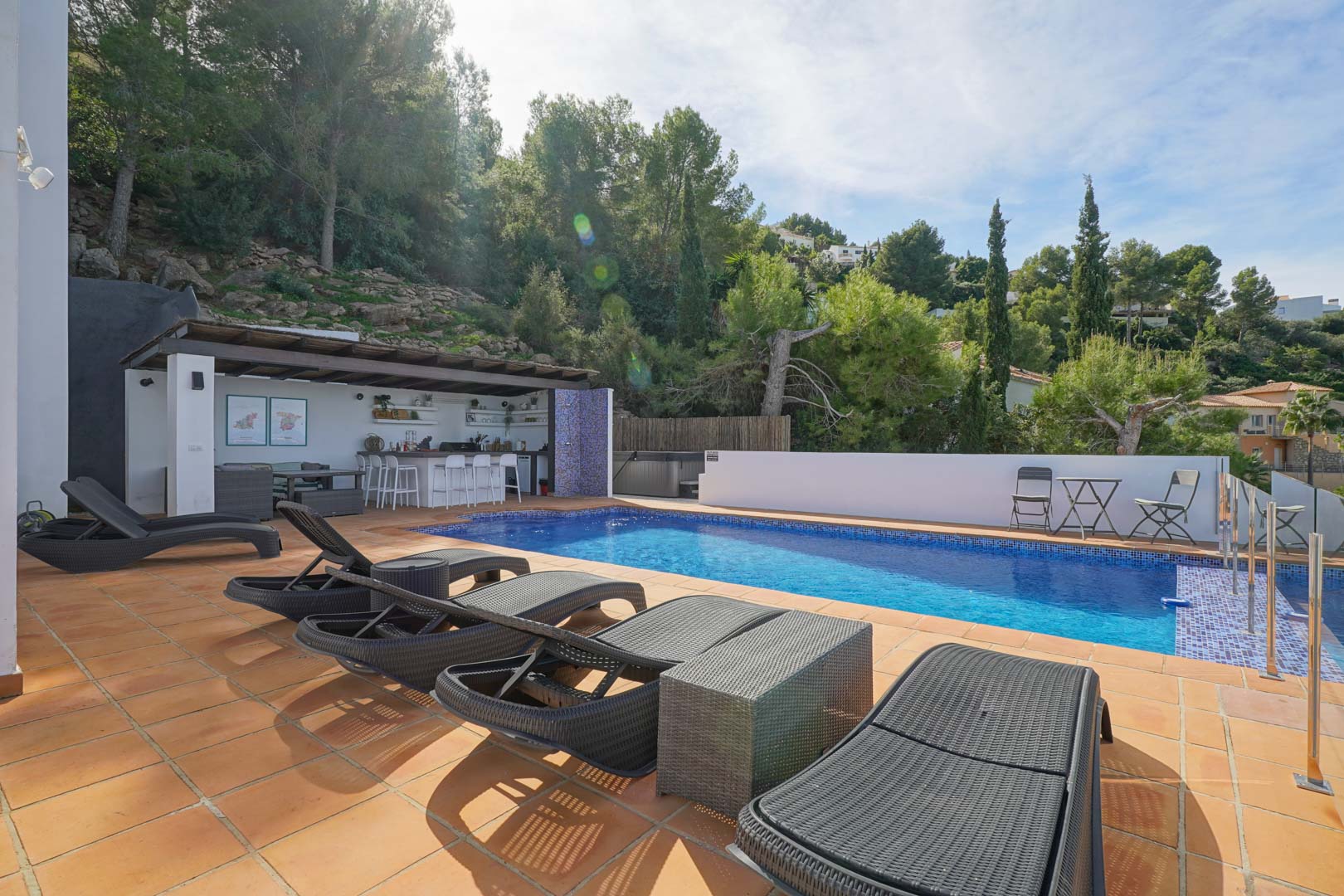 Piscina privada rodeada de una terraza de gres cerámico con tumbonas de ratán y una cocina de verano equipada bajo una pérgola de madera, junto a una ladera boscosa.
