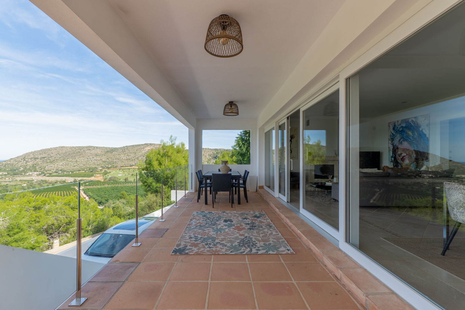 Terraza cubierta con suelo de barro, barandilla de cristal y vistas panorámicas al valle y campo de golf. Dispone de zona de comedor exterior y acceso directo al salón.