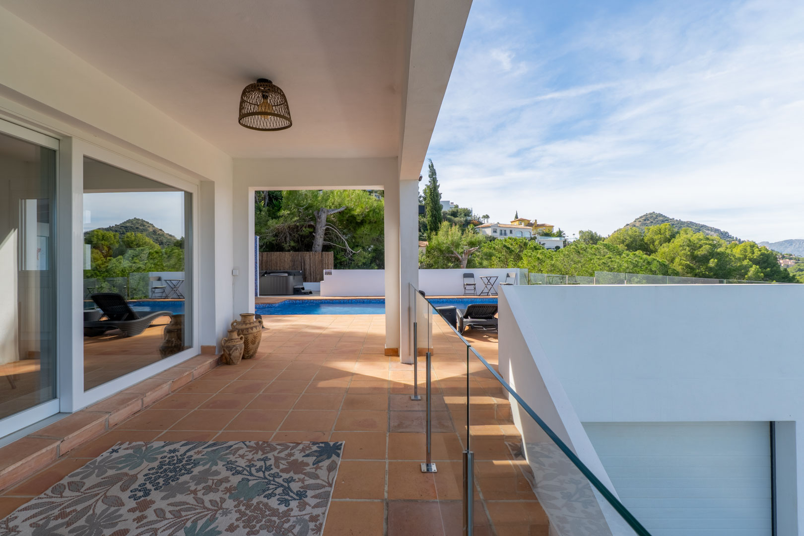 Terraza cubierta con suelos de barro, barandilla de cristal y vistas a la piscina privada. Incluye detalles decorativos como tinajas y una lámpara de mimbre.
