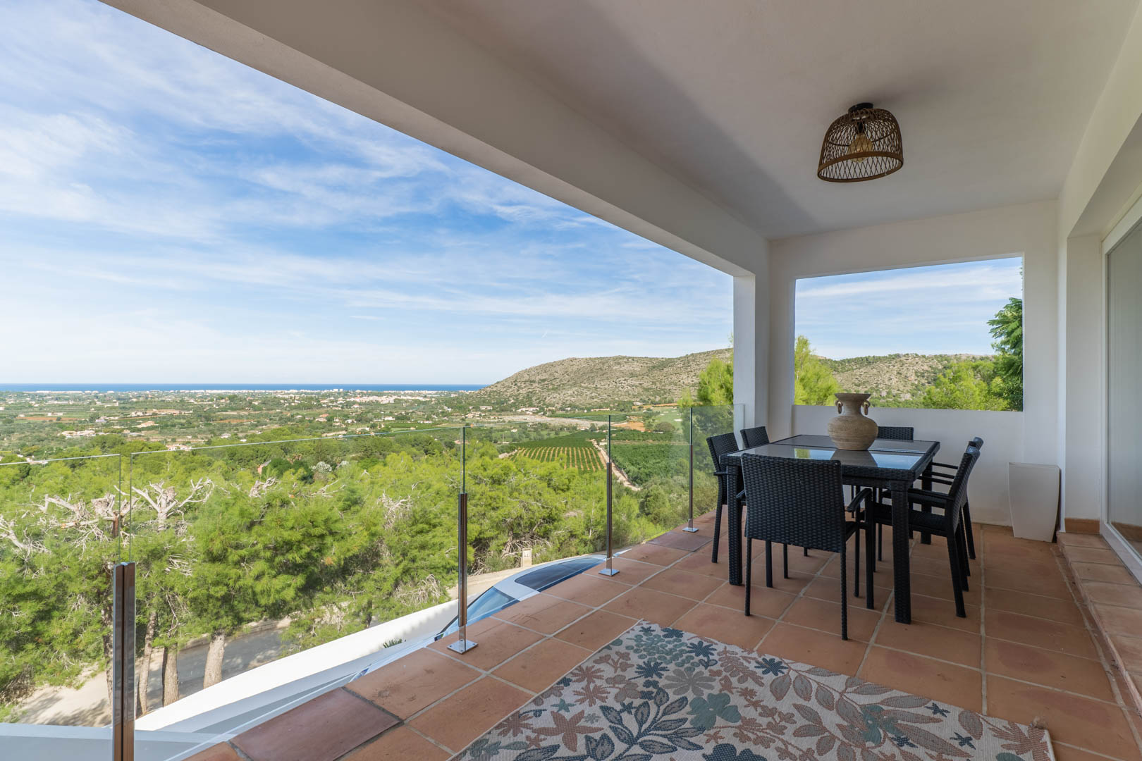 Terraza cubierta con suelos de barro, mobiliario de comedor y barandilla de cristal con vistas panorámicas al mar Mediterráneo y al valle de La Sella.