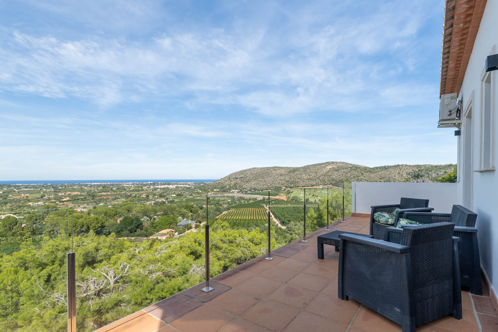 Terraza superior con barandilla de cristal, mobiliario de exterior en ratán oscuro y vistas panorámicas al mar Mediterráneo, el campo de golf y el entorno natural de La Sella.