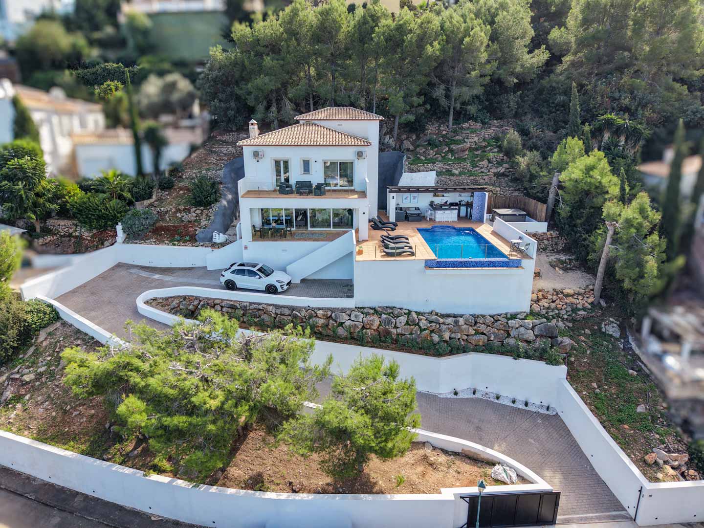 Vista aérea de la villa mediterránea con piscina infinita, cocina de verano y terrazas, integrada en una parcela elevada rodeada de pinos en La Sella.