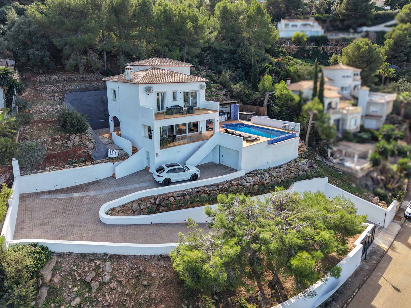 Vista exterior de la villa con fachada blanca, piscina privada, terrazas con barandillas de cristal y acceso pavimentado integrado en la ladera boscosa de La Sella.
