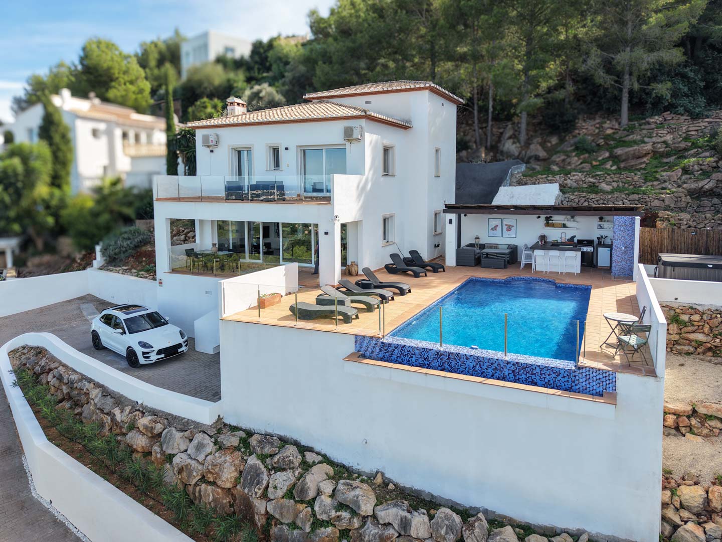 Vista exterior de la villa con piscina de gresite azul, amplia terraza con hamacas y cocina de verano equipada, rodeada de un entorno natural en La Sella.