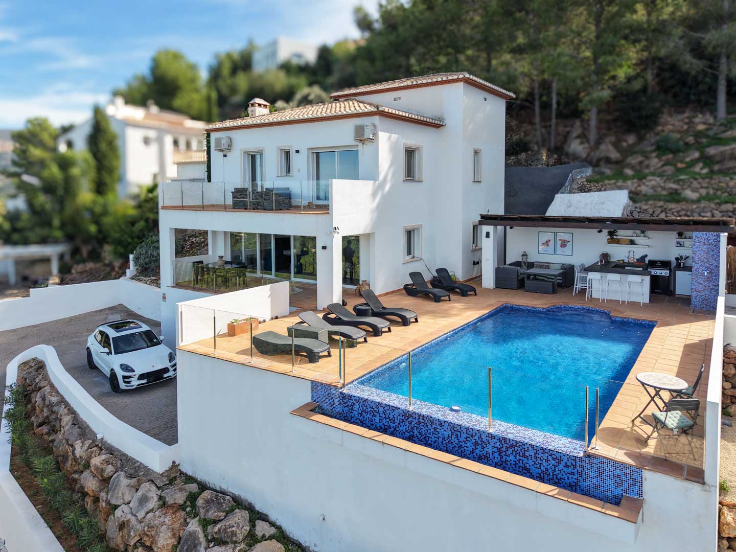 Vista exterior de la villa con piscina de mosaico azul, zona de solárium, cocina de verano bajo pérgola y fachada blanca de estilo mediterráneo integrada en la ladera.