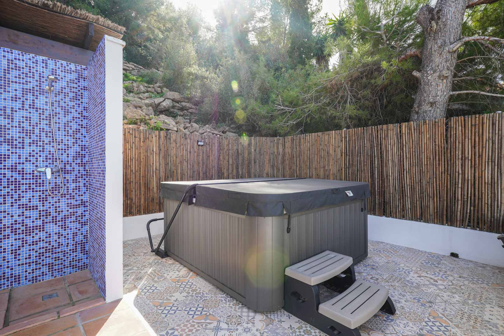 Zona exterior de bienestar con jacuzzi cubierto, ducha de mosaico azul y suelo de baldosa hidráulica, protegida por un vallado de cañizo natural.