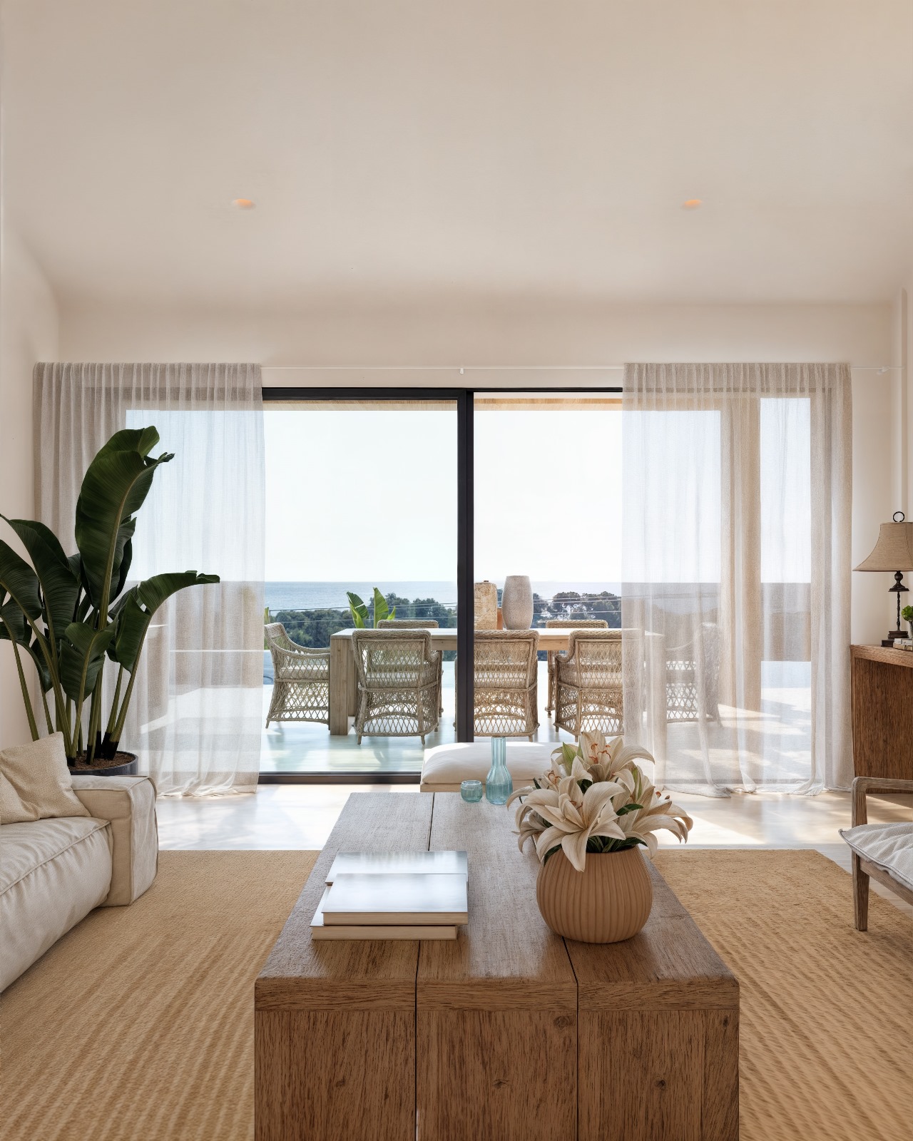 Salón de diseño ibicenco con mesa de madera natural y grandes ventanales que conectan con una terraza exterior con comedor y vistas al mar.
