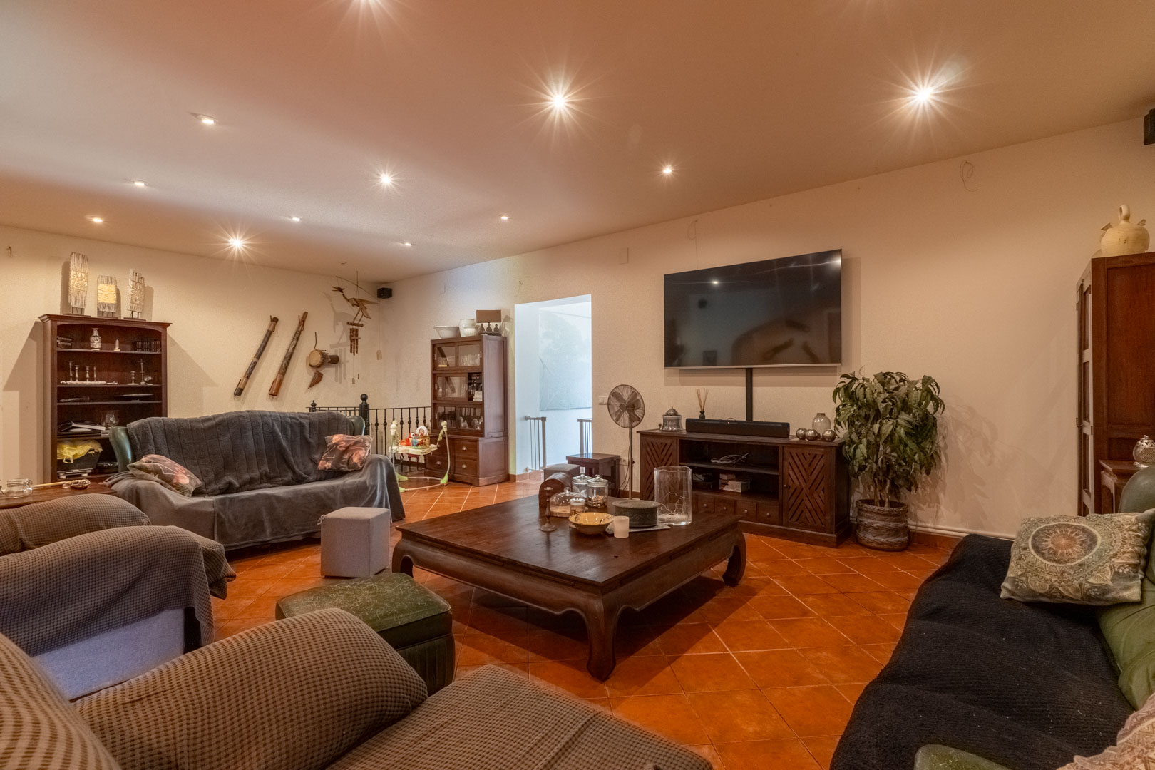 Amplia sala de estar con suelo de gres rústico, techos con iluminación empotrada y mobiliario de madera maciza. Incluye zona multimedia y acceso a la escalera con barandilla de forja.
