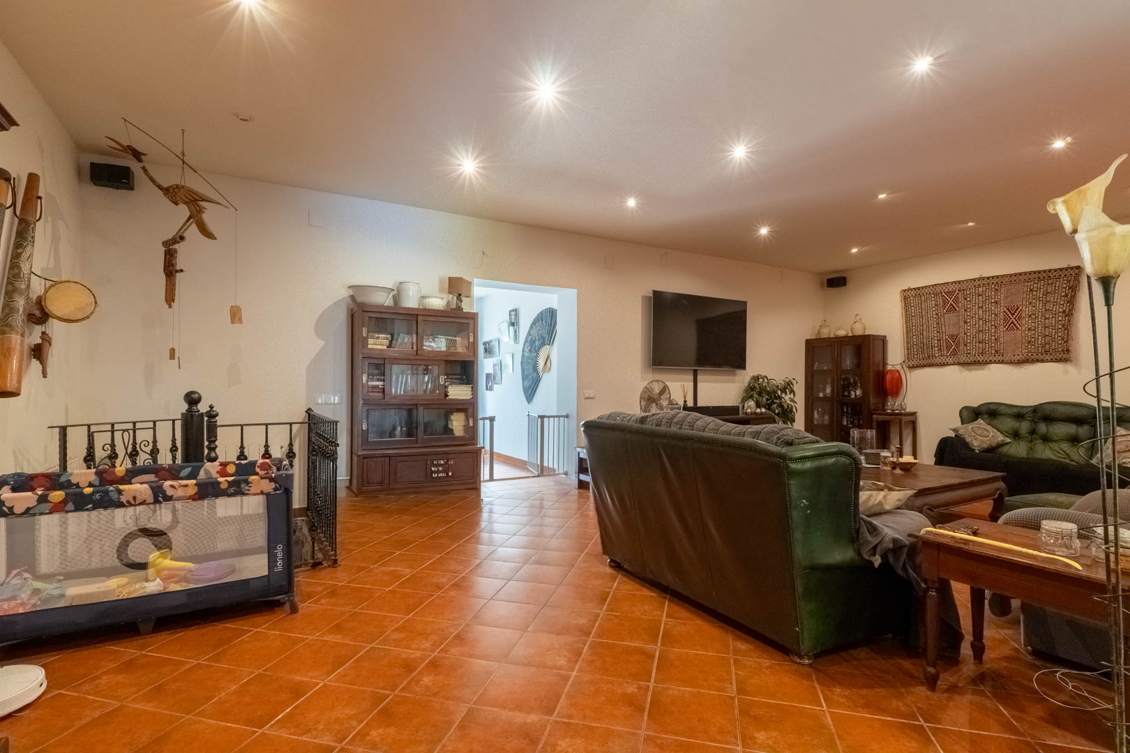 Amplio salón con suelo de baldosas de terracota, sofás de cuero verde y muebles de madera clásica. Incluye iluminación empotrada y acceso a la planta inferior mediante escalera de forja.