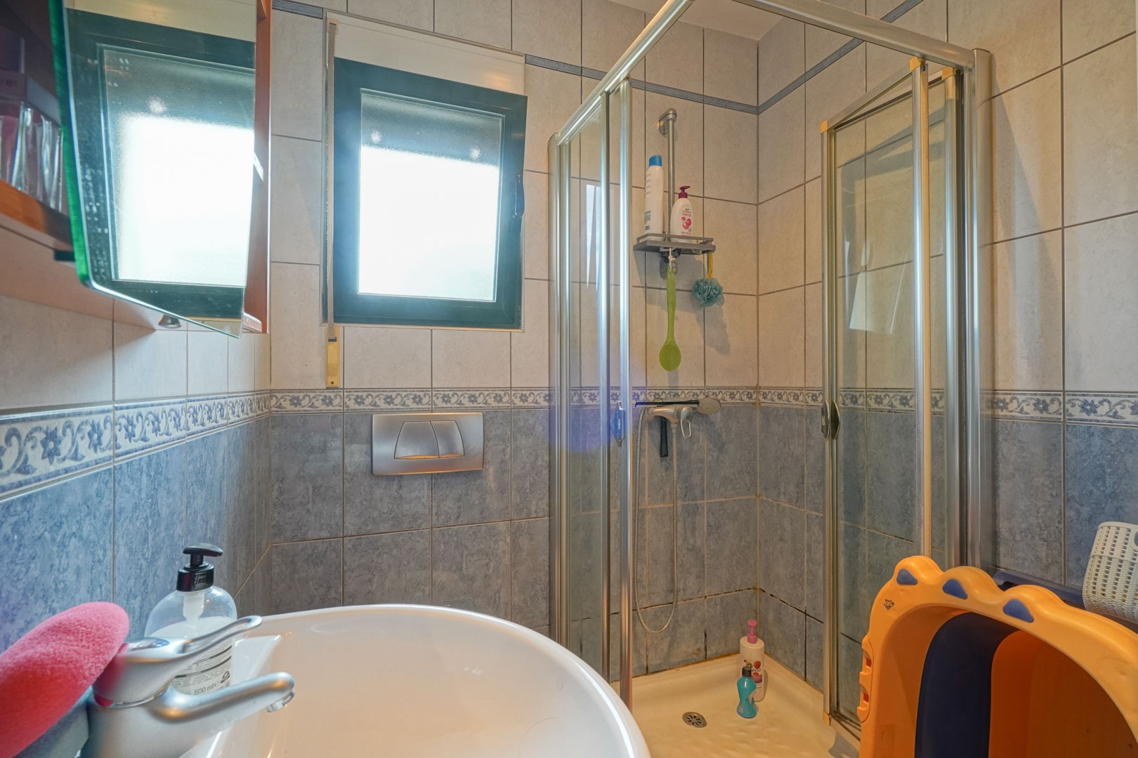 Baño funcional con ducha de esquina con mampara de cristal, lavabo cerámico blanco y paredes alicatadas en tonos azules con cenefa decorativa y ventana oscilobatiente.