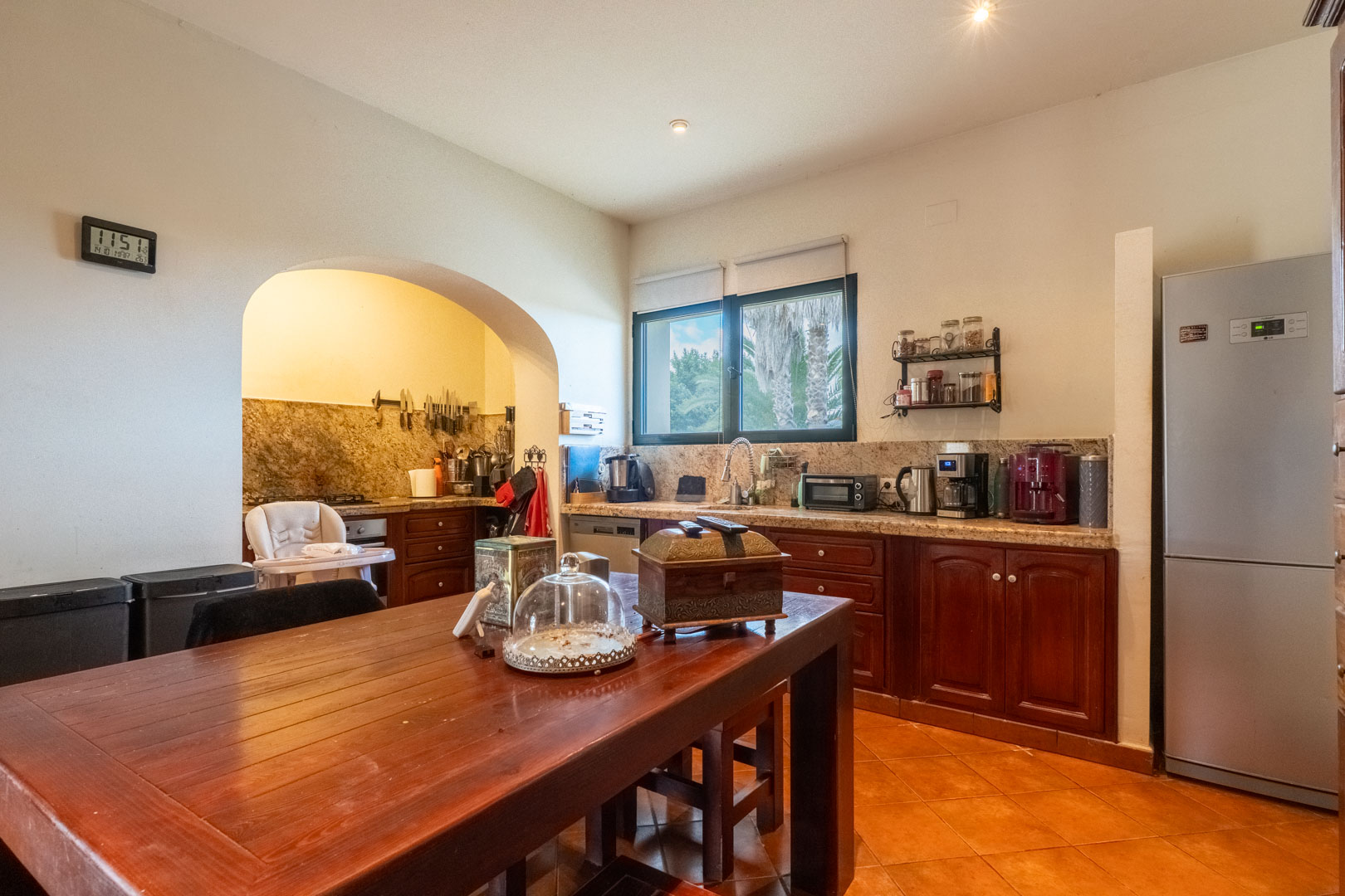 Cocina rústica con armarios de madera oscura, encimeras de granito y mesa de comedor central. Un arco conecta con la zona de cocción sobre un suelo de baldosas de terracota.