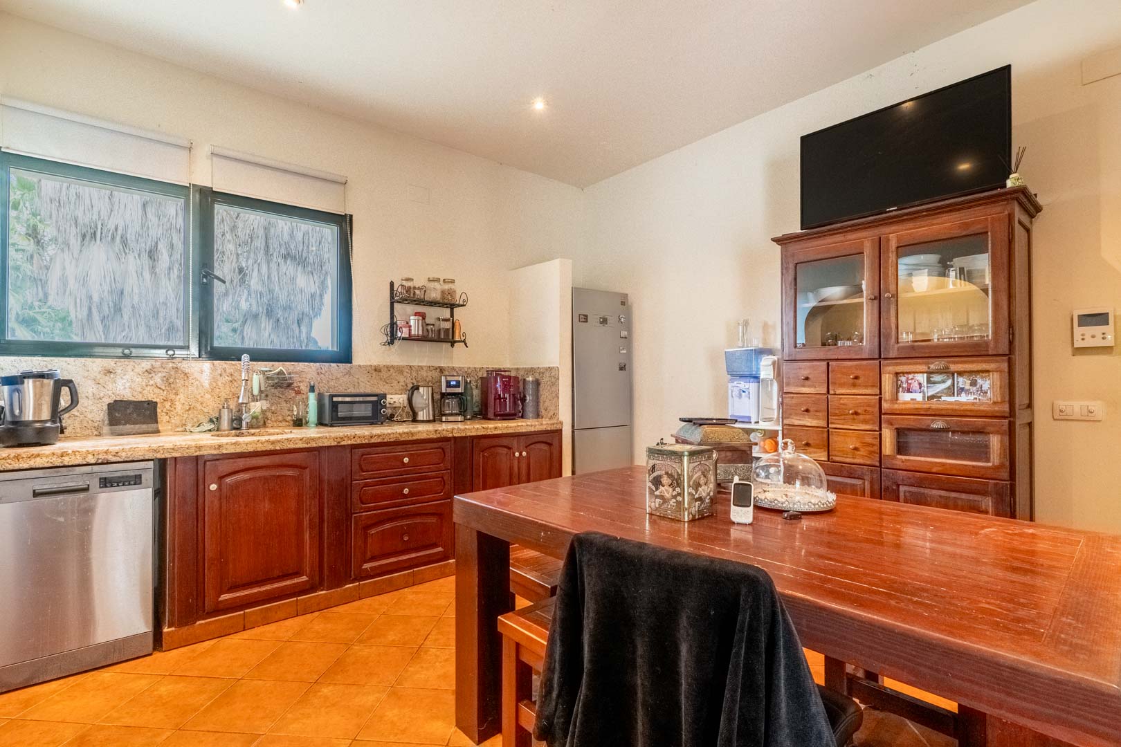 Cocina rústica con encimera de granito, muebles de madera oscura y mesa de comedor central. Dispone de electrodomésticos de acero inoxidable y luz natural a través del gran ventanal.
