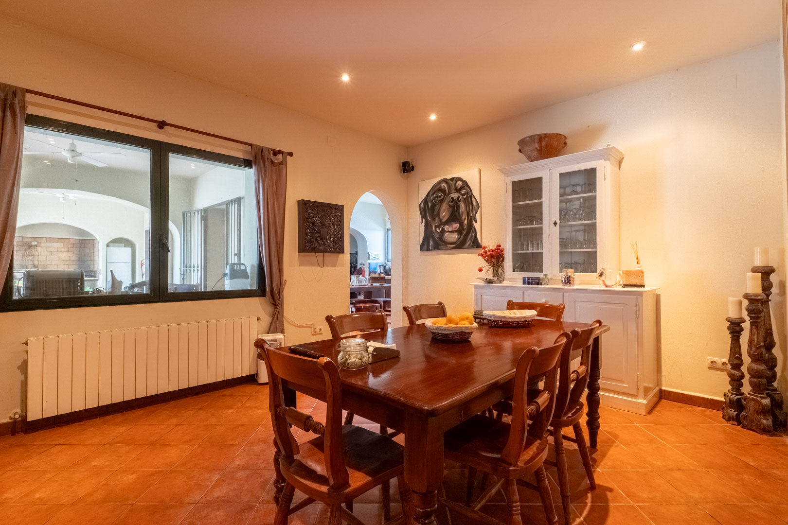 Comedor con suelo de terracota, mesa de madera maciza y aparador blanco. Incluye radiador bajo la ventana y acceso mediante arco a la estancia contigua.