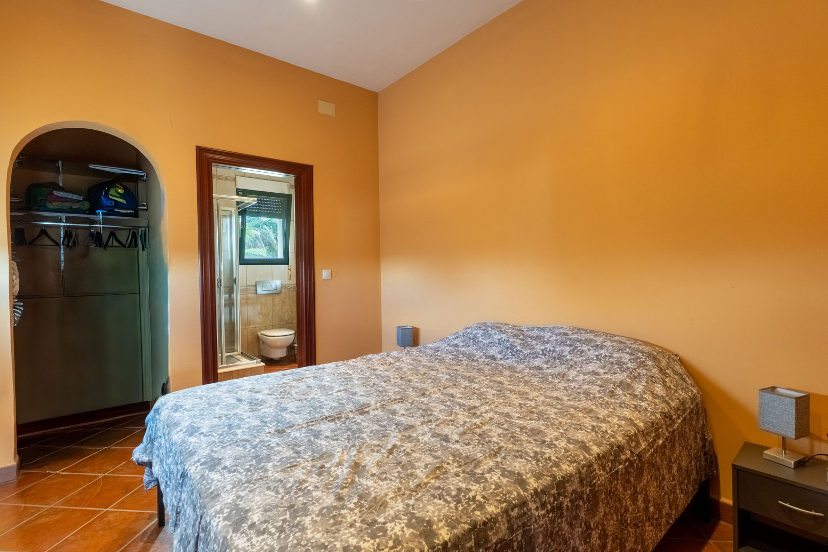 Dormitorio con suelos de terracota y paredes en tonos cálidos, que incluye un vestidor con arco y baño en suite con plato de ducha.