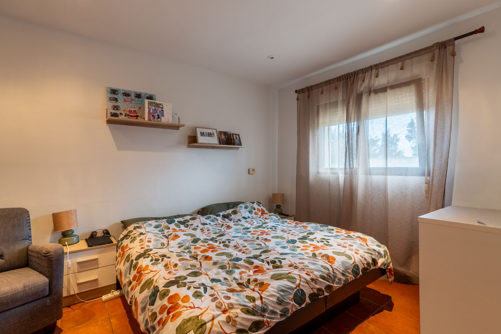 Dormitorio luminoso con suelo de terracota, paredes blancas y estantes de madera. Incluye una ventana con cortinas traslúcidas y persiana que aporta luz natural.