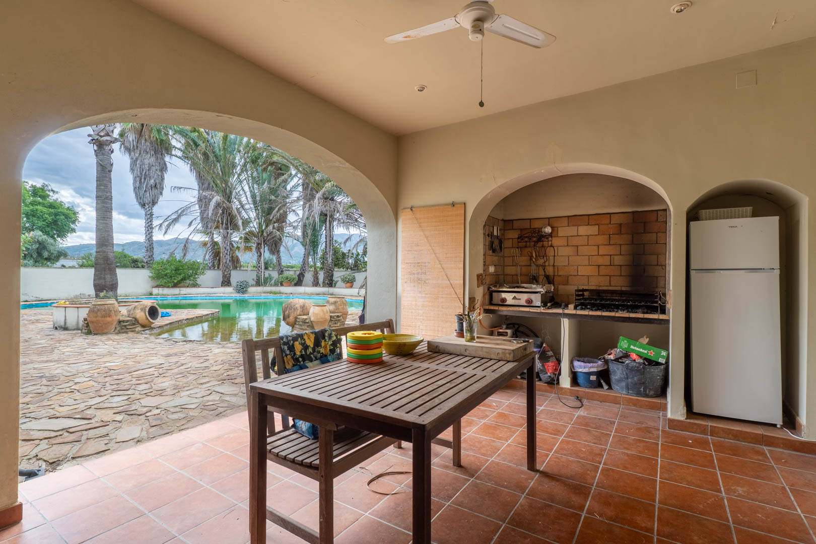 Porche cubierto con zona de barbacoa de obra, mesa de comedor de madera y vistas directas a la piscina y jardines a través de un gran arco mediterráneo.