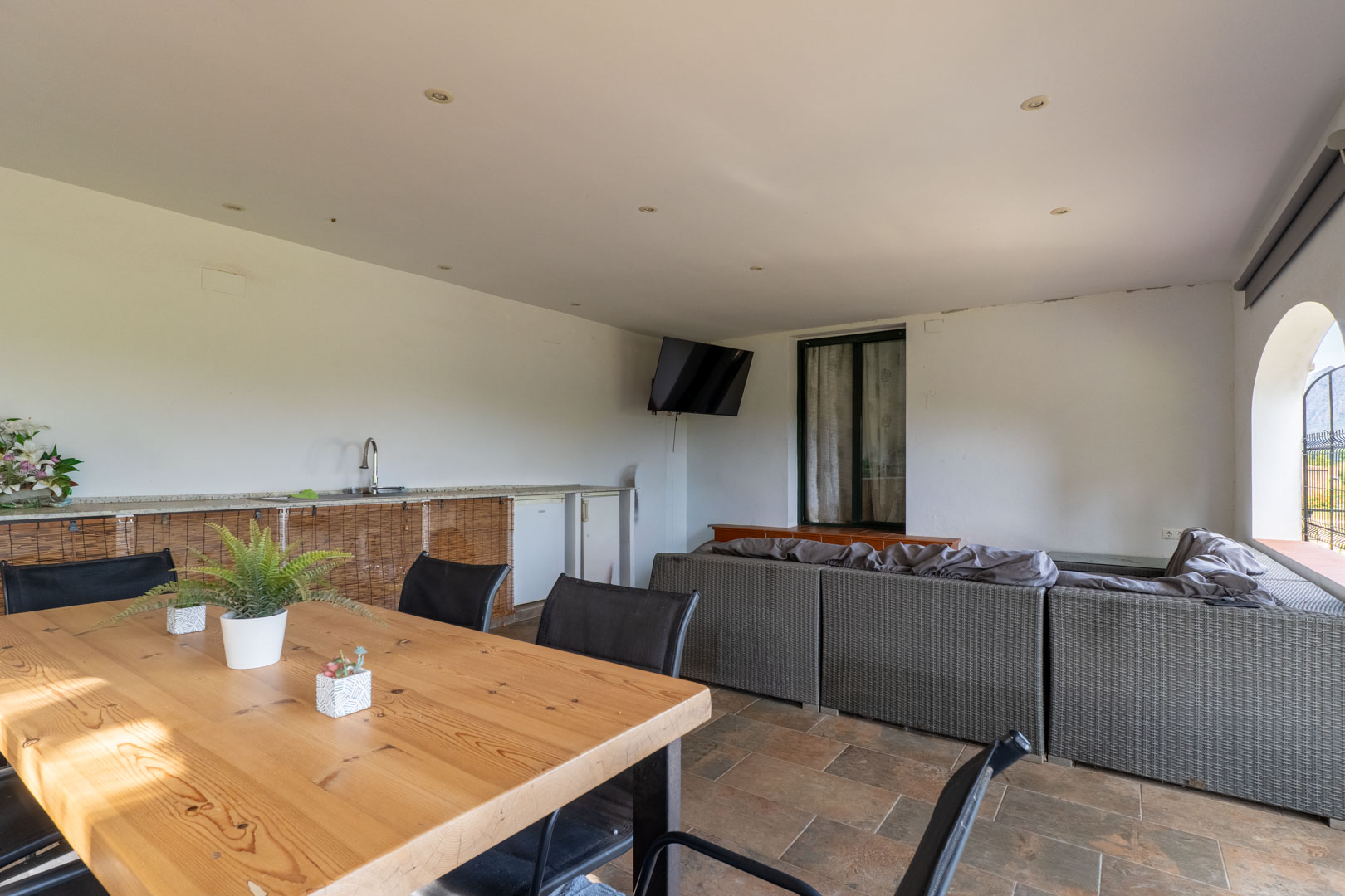 Porche cubierto con zona de estar de mimbre, mesa de comedor de madera maciza y cocina exterior integrada. Incluye iluminación empotrada y suelos cerámicos de tono natural.