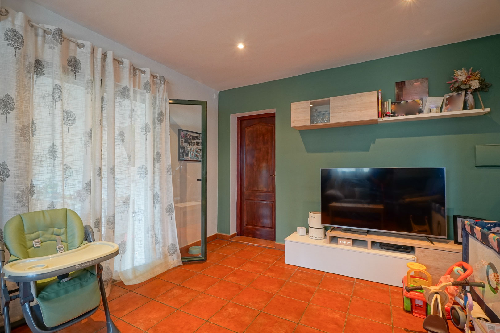 Sala de estar con pared verde salvia, suelo de baldosas de terracota y mobiliario funcional. Incluye acceso mediante puerta de madera y luz natural filtrada por cortinas.