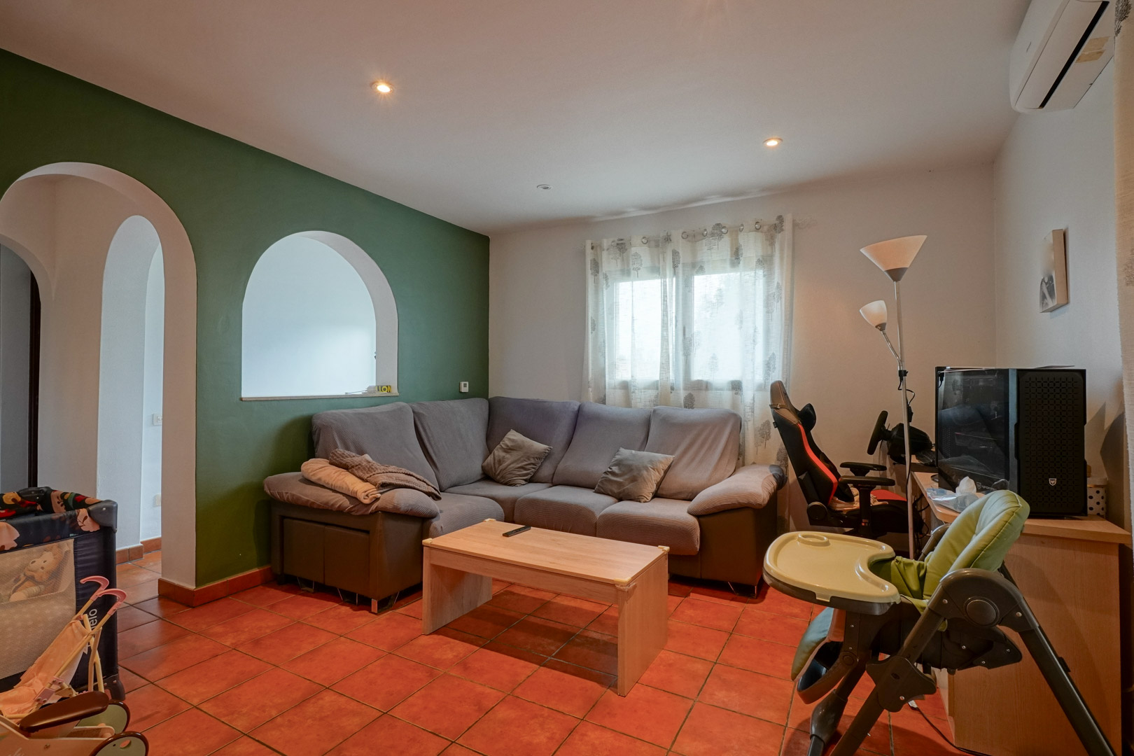Sala de estar con suelo de terracota, pared de acento verde y arcos decorativos. Incluye sofá rinconero, zona de despacho y aire acondicionado tipo split.