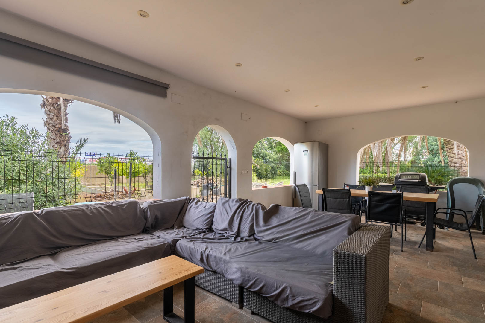 Terraza cubierta con arcos mediterráneos, zona de estar con sofás grises, comedor exterior y cocina equipada con barbacoa. Suelo cerámico y vistas al jardín.