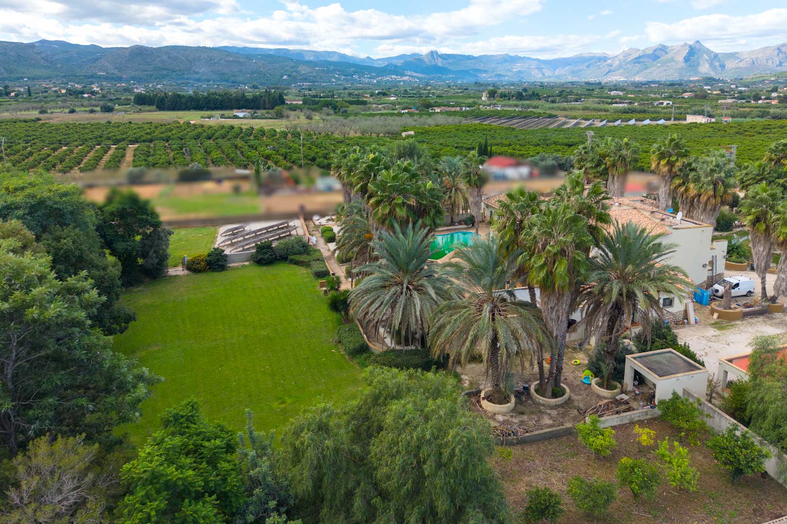 Vista aérea de la extensa finca con jardines de palmeras, zona de césped y piscina, rodeada de campos agrícolas y vistas a las montañas de Ondara.