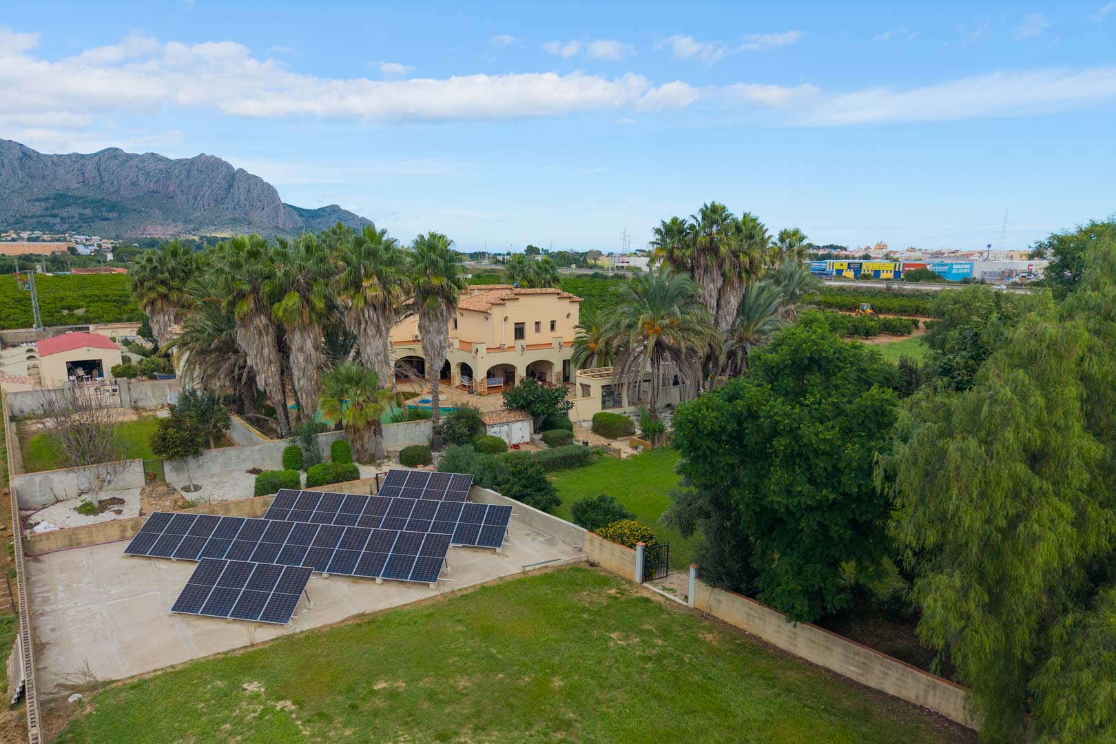 Vista aérea de la finca con instalación de placas solares, jardines con palmeras y la villa principal con arcos mediterráneos frente a la montaña.