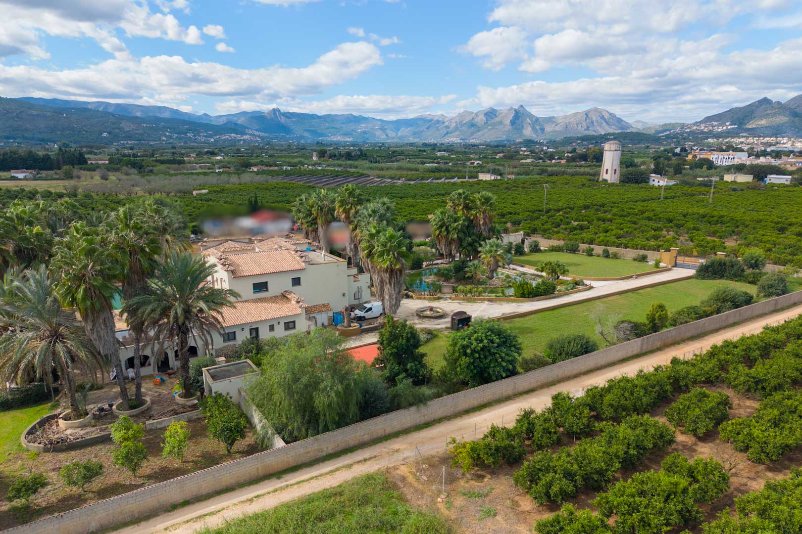 Vista aérea de la finca que muestra la casa principal, jardines con palmeras y un estanque, rodeada de extensos campos y con vistas panorámicas a las montañas.