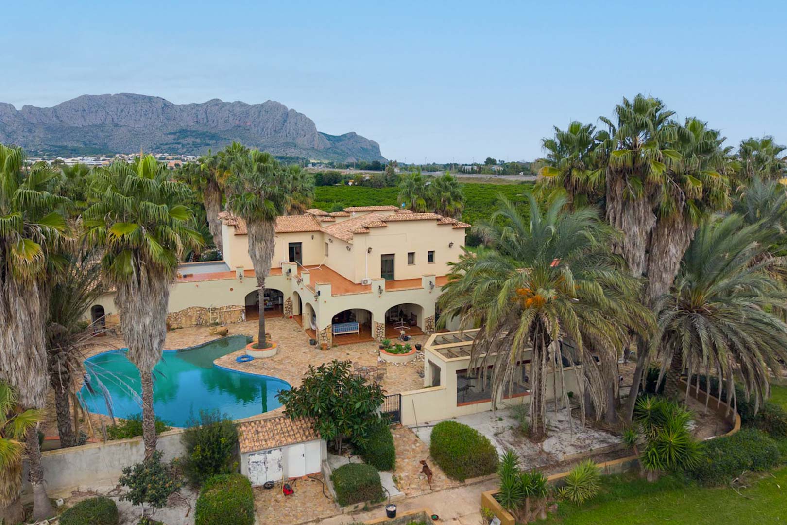 Vista aérea de la villa con piscina de diseño orgánico, terrazas porticadas y frondosos jardines de palmeras bajo la silueta de la Sierra de Segaria.