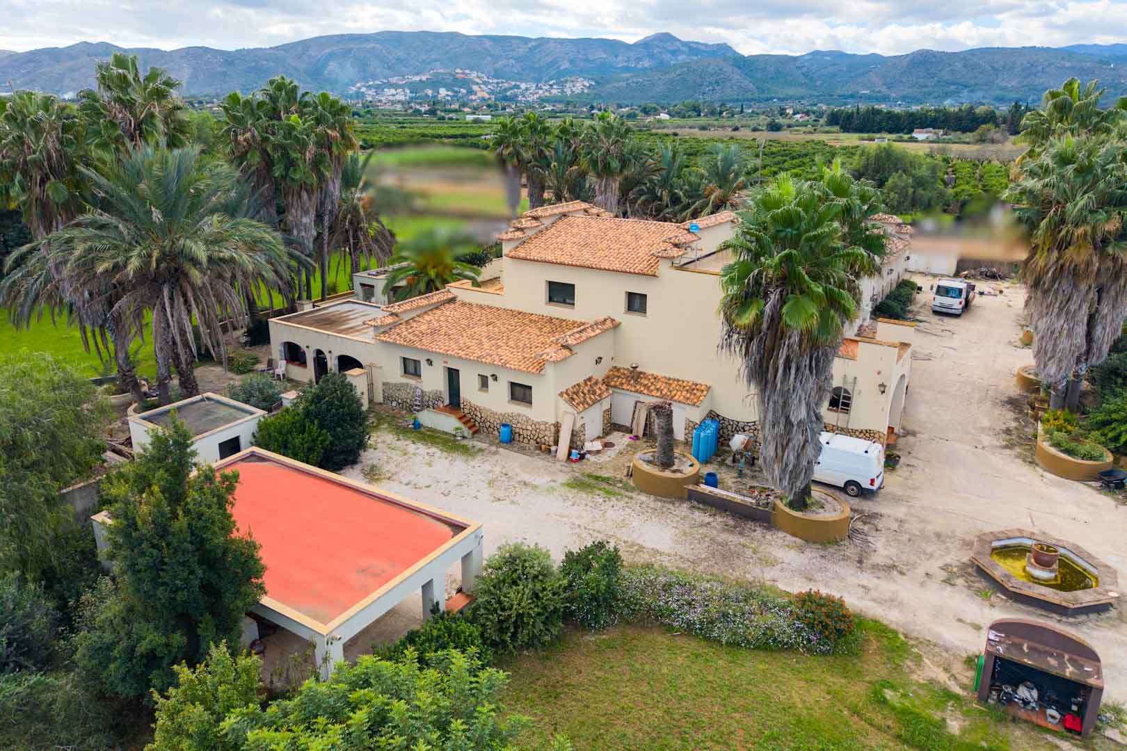 Vista aérea de la villa mediterránea con tejados de terracota, rodeada de palmeras y jardines, con vistas panorámicas al valle y las montañas de Ondara.
