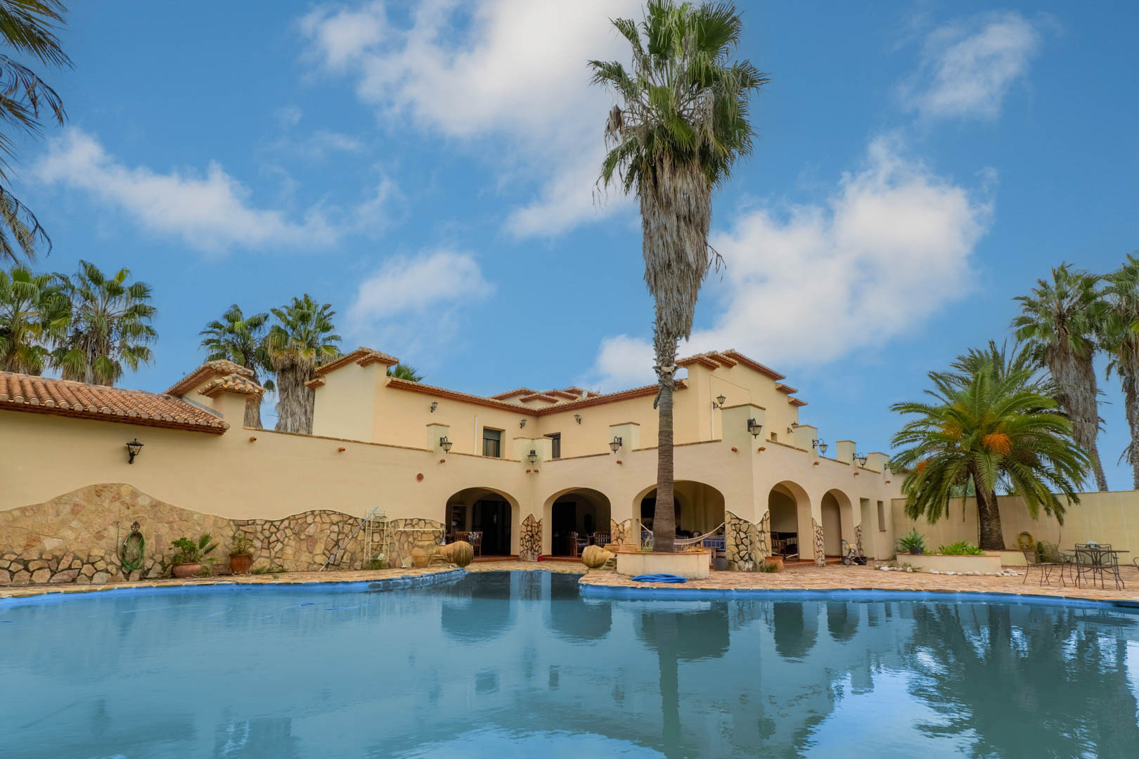 Vista exterior de la piscina de forma irregular rodeada de una terraza de piedra, con la fachada mediterránea de la villa y sus porches arqueados al fondo.