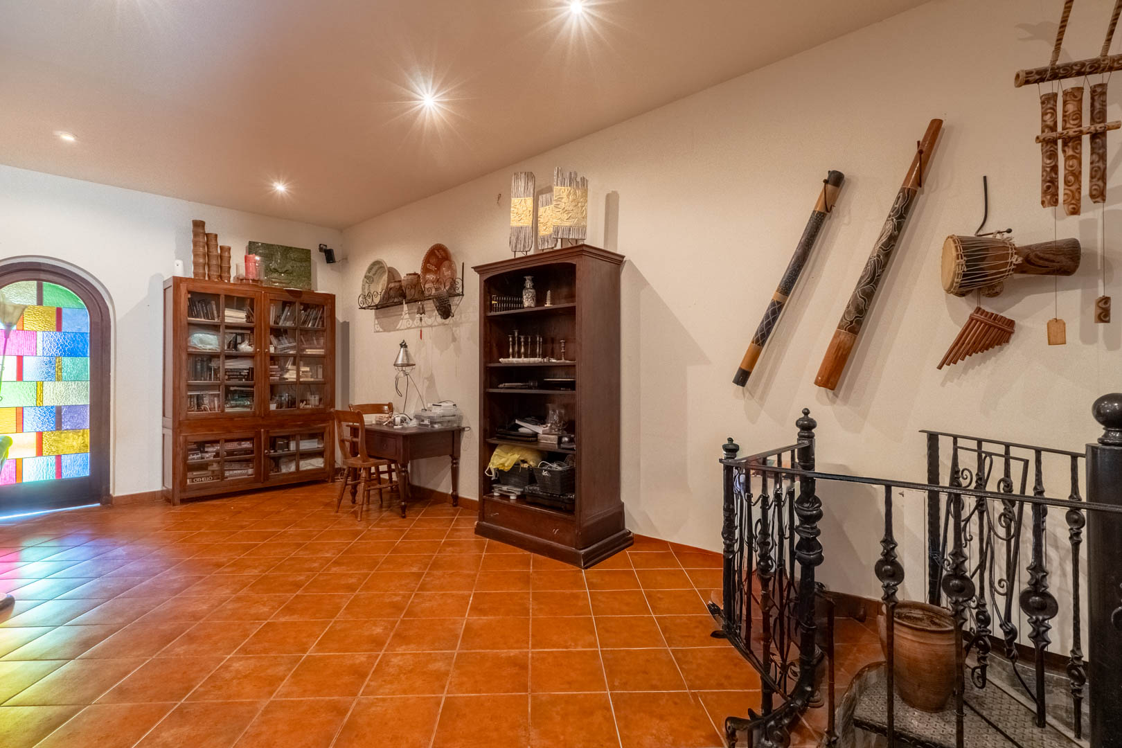 Zona de estudio con suelos de terracota, librerías de madera, instrumentos étnicos decorativos y acceso a la escalera de caracol con barandilla de hierro.
