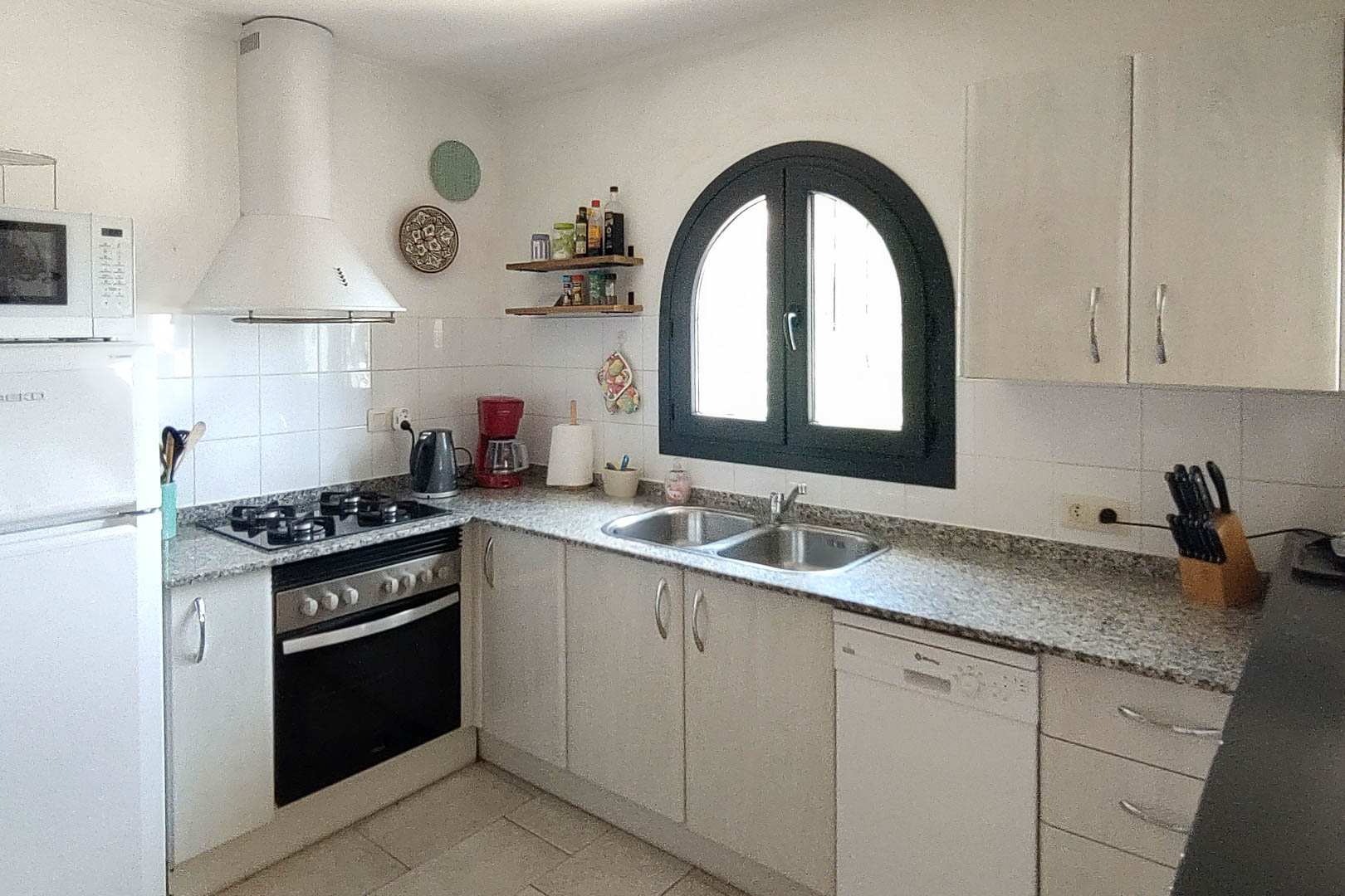 Cocina funcional con encimera de granito, equipada con horno, placa de gas y lavavajillas. Destaca su ventana de arco con marco oscuro que aporta luz natural a la zona de trabajo.