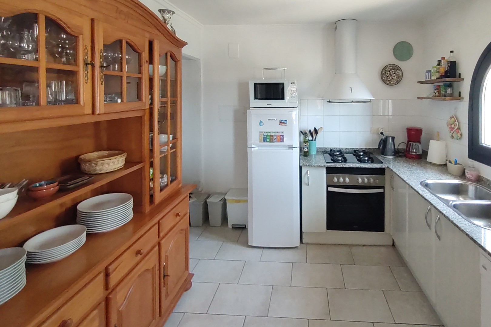 Cocina funcional equipada con encimera de granito, fogones de gas y un amplio aparador de madera maciza para vajilla, sobre un suelo de baldosa clara.