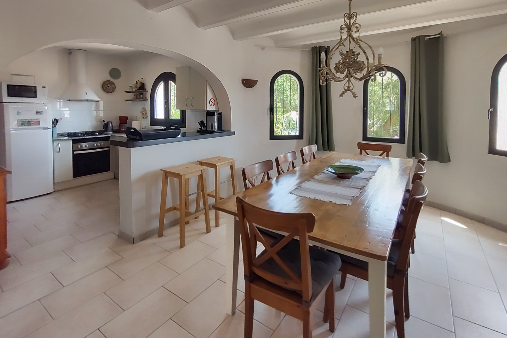 Comedor con mesa de madera para ocho comensales y cocina abierta tras un arco, equipada con barra, taburetes y electrodomésticos bajo ventanas de medio punto.