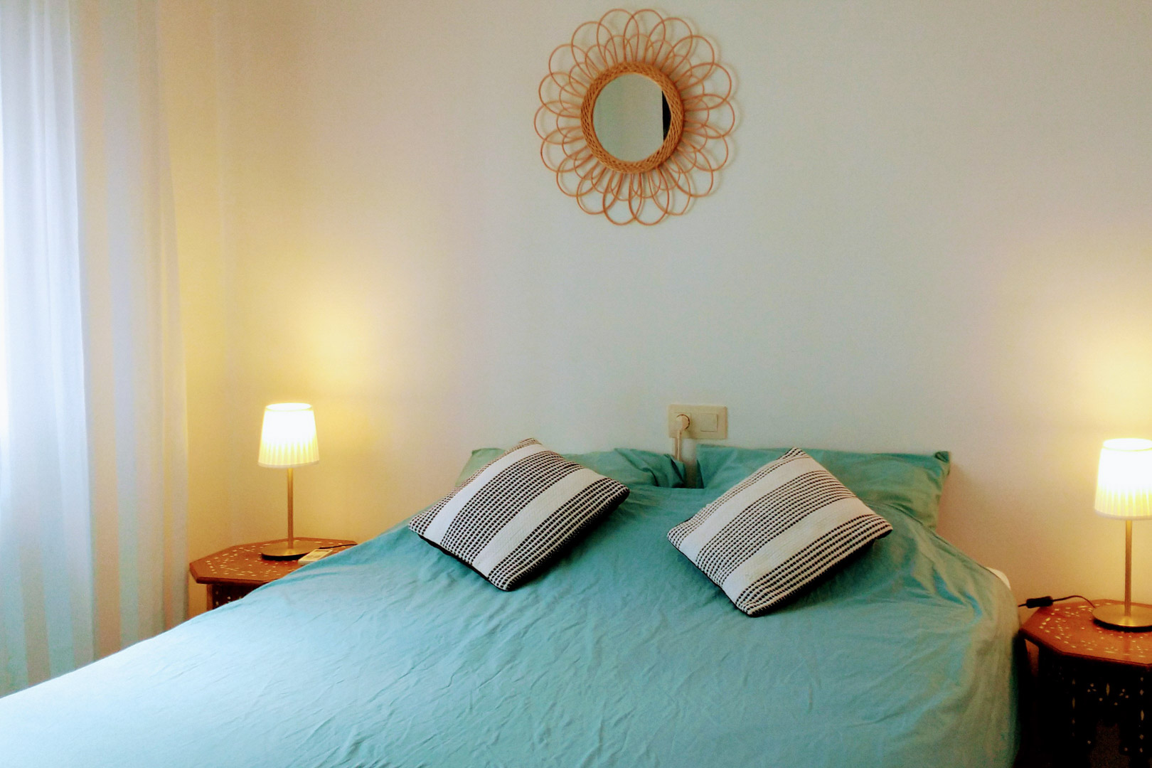 Dormitorio con cama doble, ropa de cama turquesa, mesitas de noche hexagonales de madera y un espejo decorativo de ratán sobre la pared blanca.