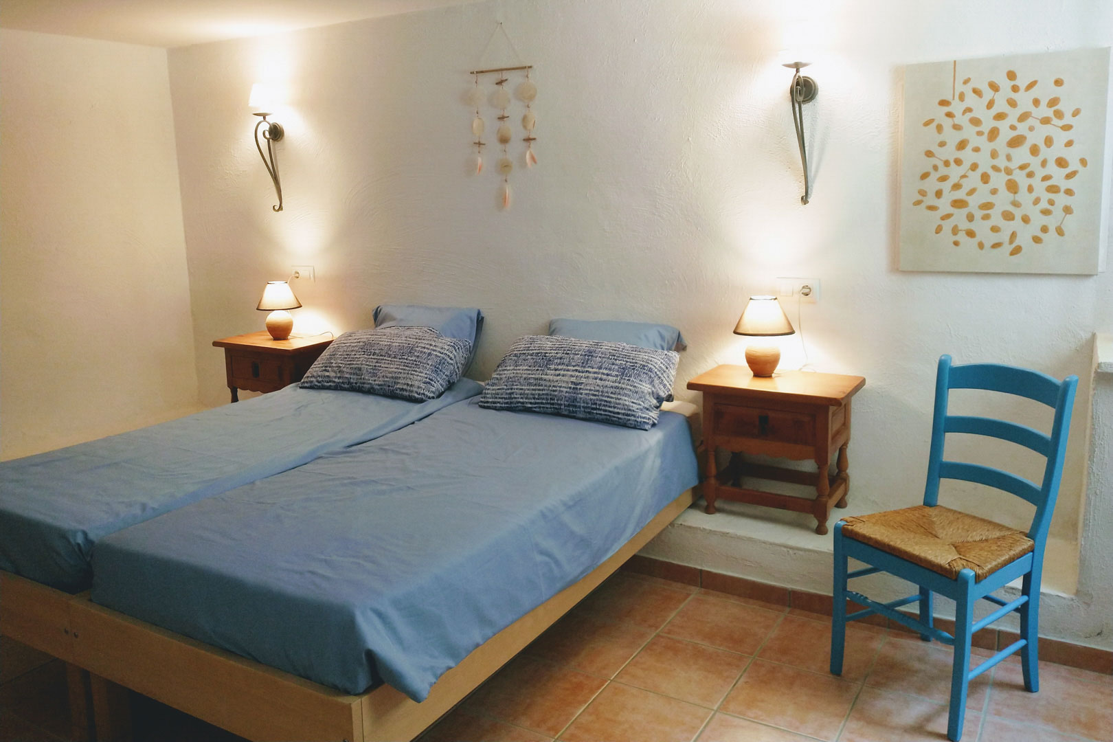 Dormitorio con dos camas individuales, suelos de terracota y paredes blancas. Incluye mesitas de madera, iluminación cálida y una silla rústica azul.