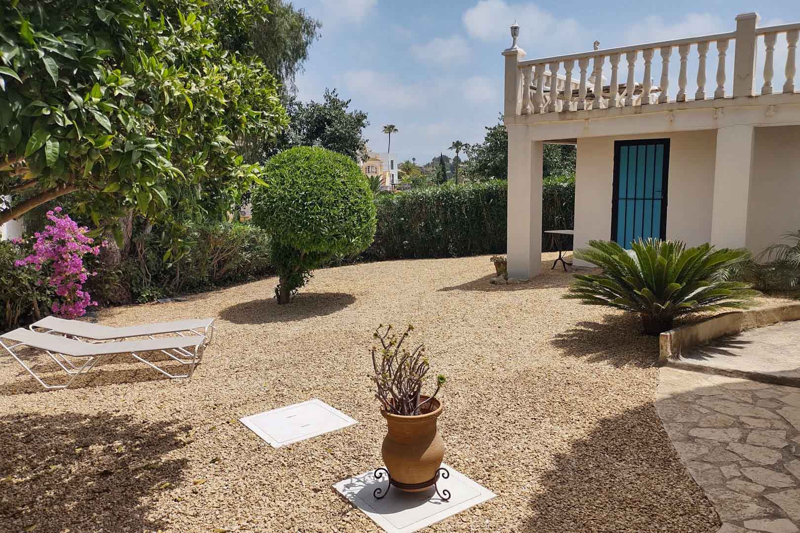 Jardín mediterráneo de bajo mantenimiento con zona de grava, tumbonas, vegetación autóctona y fachada blanca con balaustrada clásica y puerta azul.