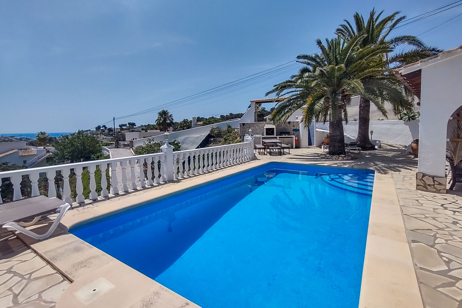 Piscina exterior con terraza de piedra, balaustrada blanca y vistas al mar. Al fondo, zona de barbacoa bajo palmeras y comedor al aire libre.