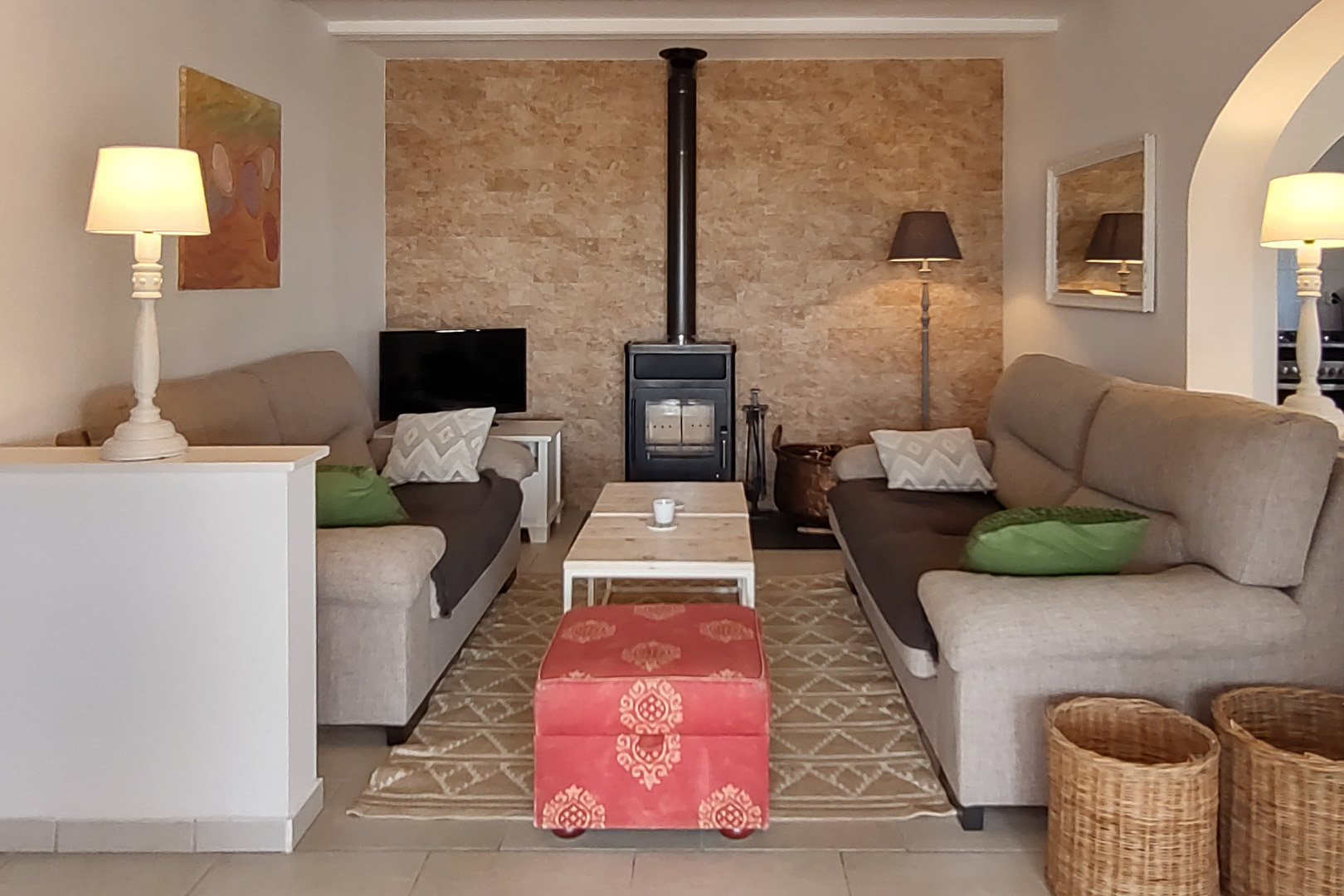 Salón acogedor con estufa de leña sobre pared de piedra natural, sofás enfrentados, mesa de centro de madera y un puf rojo decorativo en un ambiente cálido.