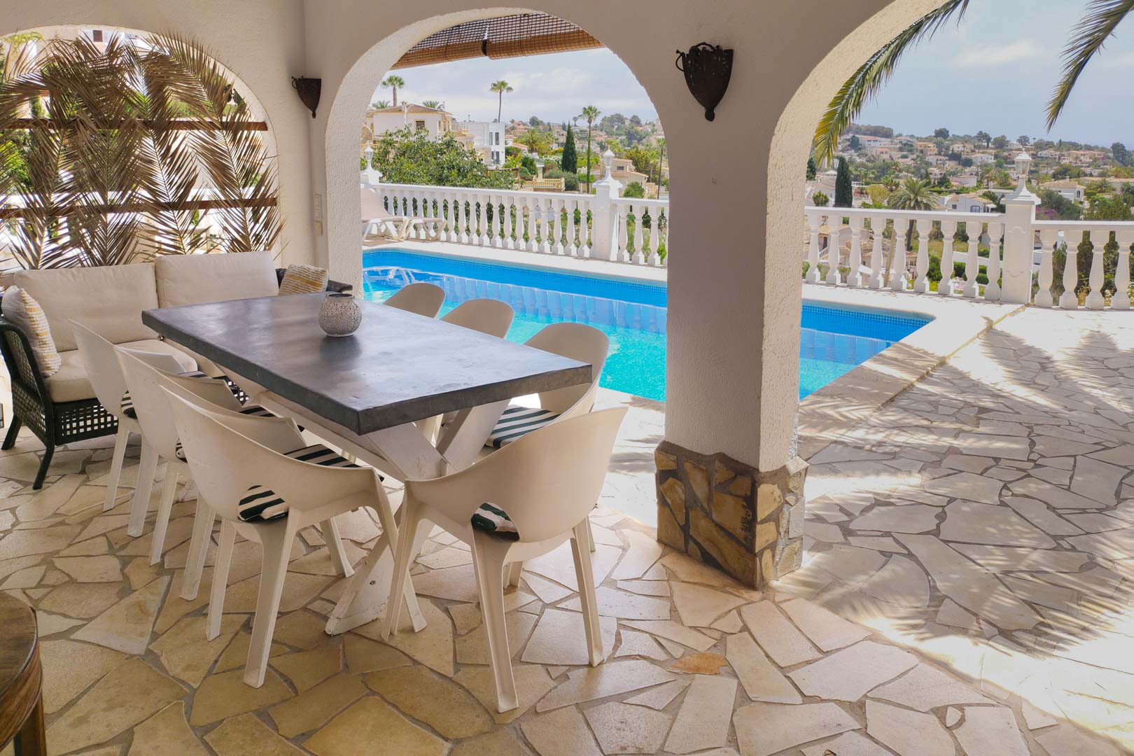 Terraza cubierta con arcos tradicionales, suelo de piedra natural y zona de comedor frente a la piscina con vistas despejadas al valle y palmeras.