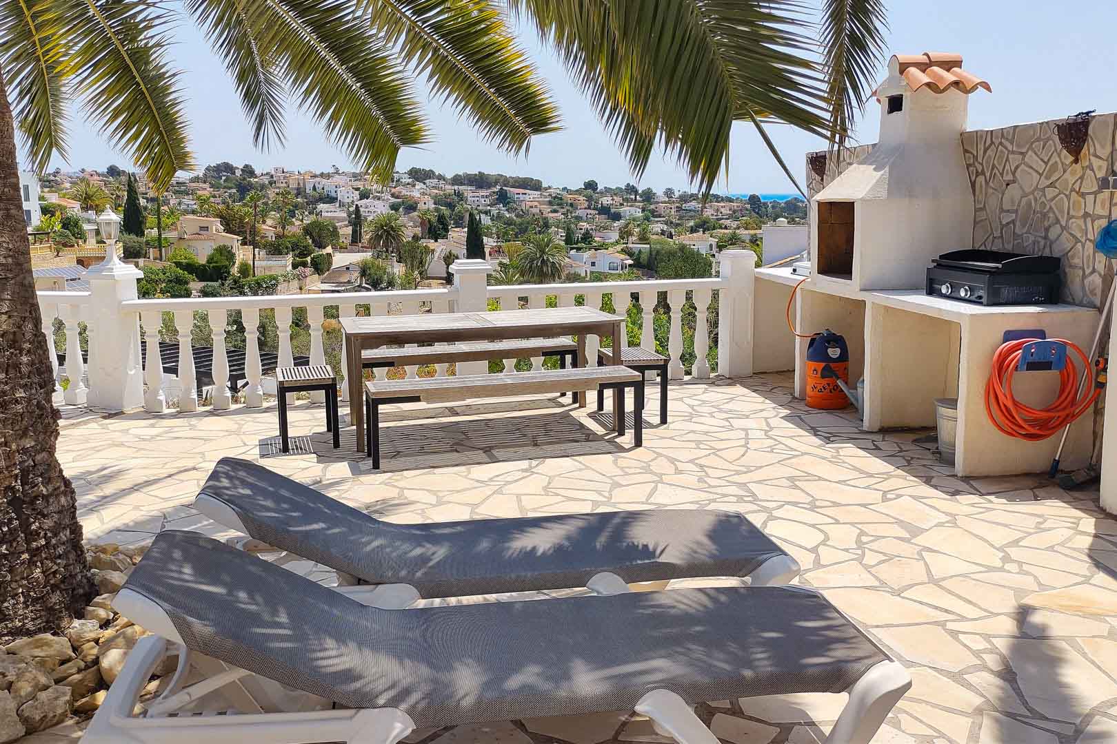 Terraza exterior pavimentada en piedra con zona de barbacoa de obra, mesa de picnic de madera y tumbonas bajo la sombra de palmeras con vistas al valle.