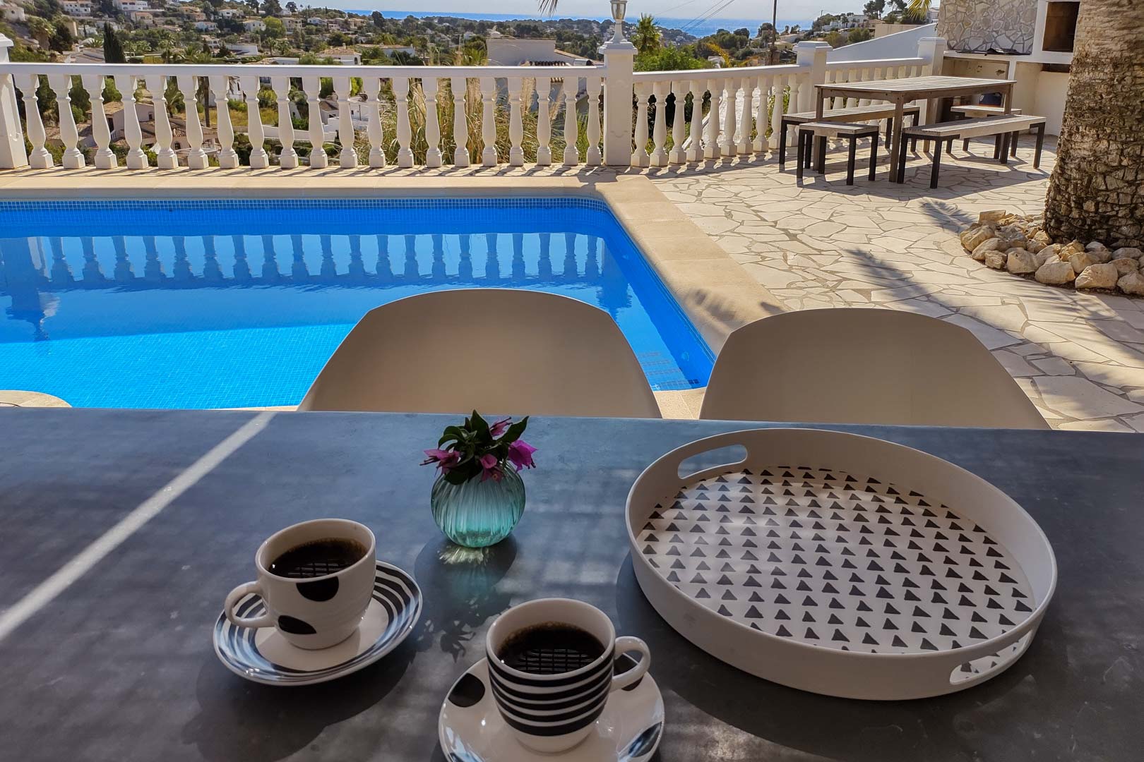 Terraza principal con vistas al mar y piscina privada. Incluye zona de comedor exterior con mesa de madera y bancos sobre suelo de piedra natural y balustrada blanca.