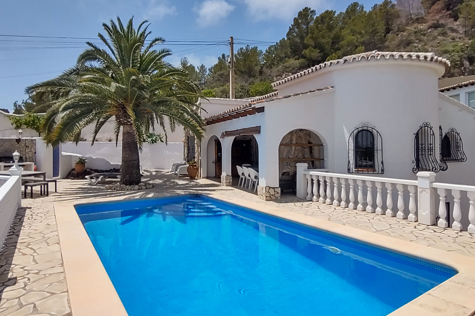 Vista de la piscina rectangular y terraza de piedra natural, destacando la arquitectura blanca con arcos, rejas de hierro y una gran palmera bajo el cielo despejado.