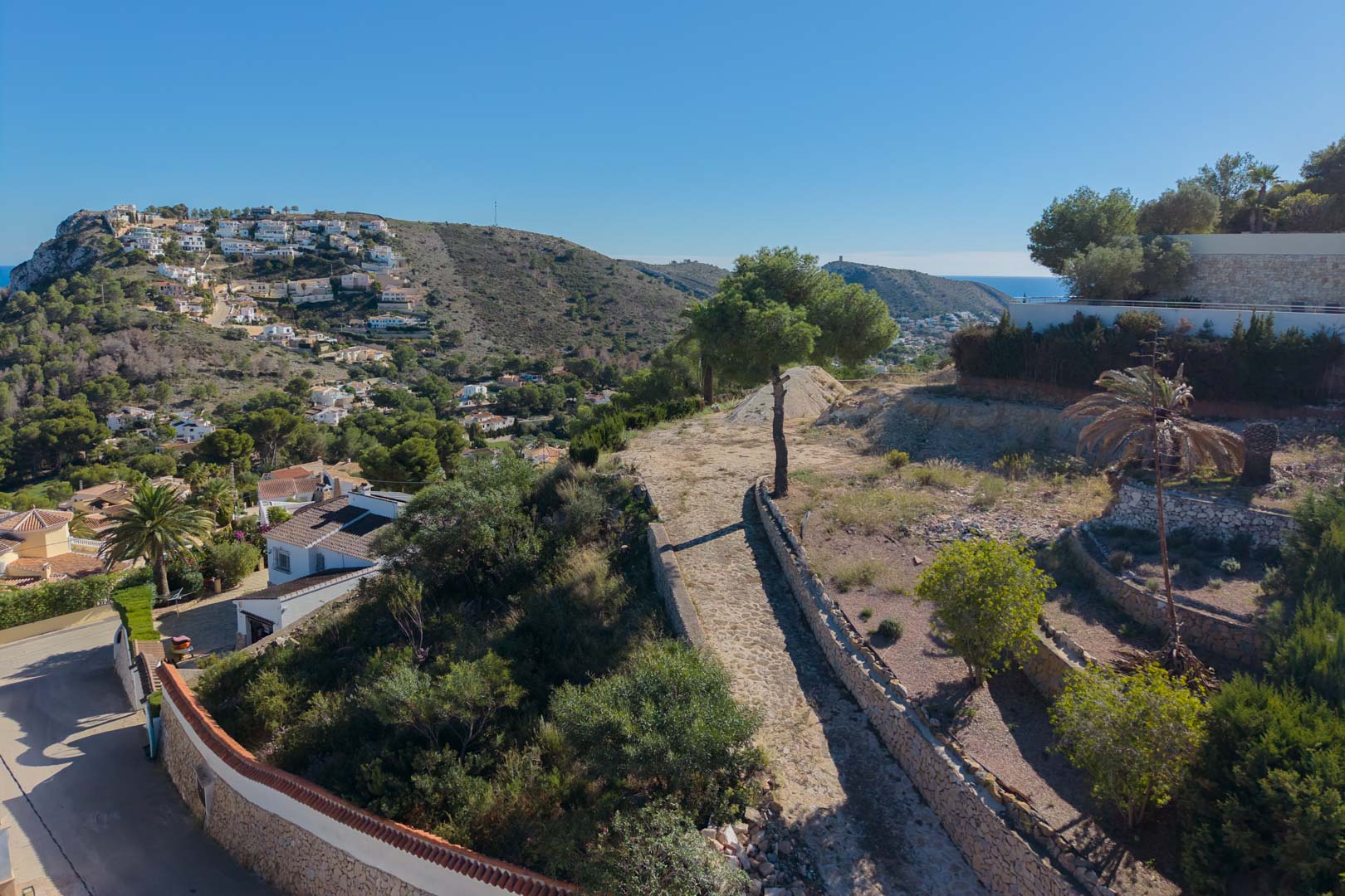 Vista elevada de la parcela con muros de mampostería de piedra natural y vegetación mediterránea, ofreciendo vistas panorámicas al valle y al mar en Moraira.