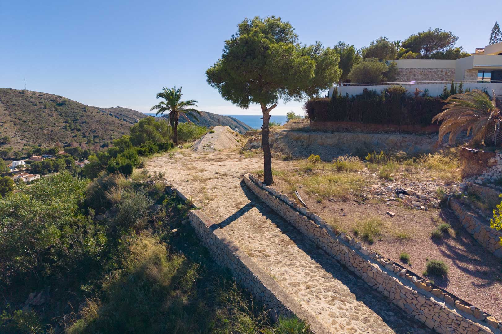 Vista panorámica desde la parcela con camino empedrado y muros de piedra seca, rodeada de vegetación mediterránea y vistas despejadas al mar.